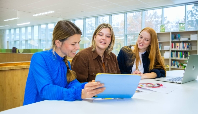 Drie mensen zitten aan een tafel met een tablet en een laptop en lachen.