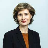 Portrait headshot. The picture is taken in front of a blue background and the subject is wearing a black blazer with an orange shirt underneath. Her hair is brunette with blonde highlights and is chin length, and her eyes are brown. She is facing the camera, with shoulders slightly towards the right.