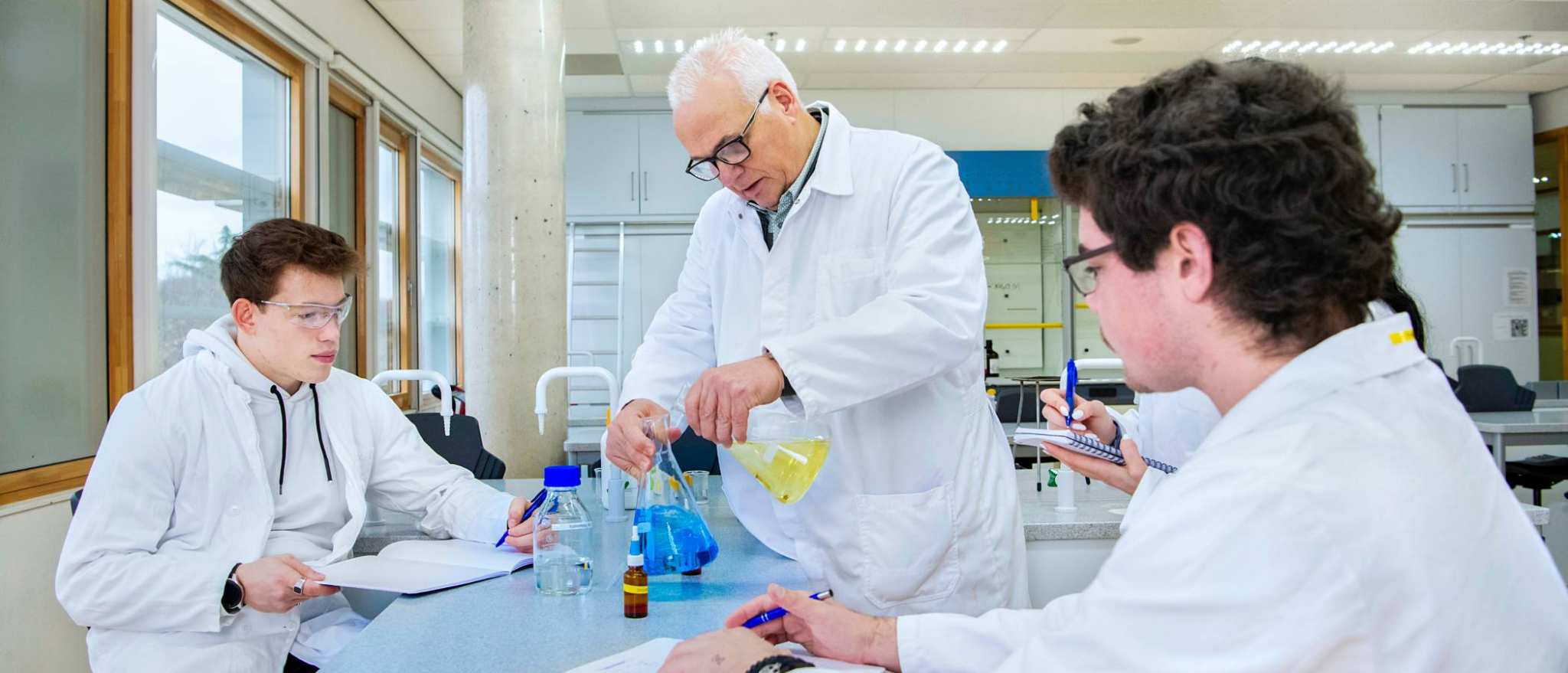 De docent doet twee vloeistoffen bij elkaar, waarbij drie studenten toekijken.