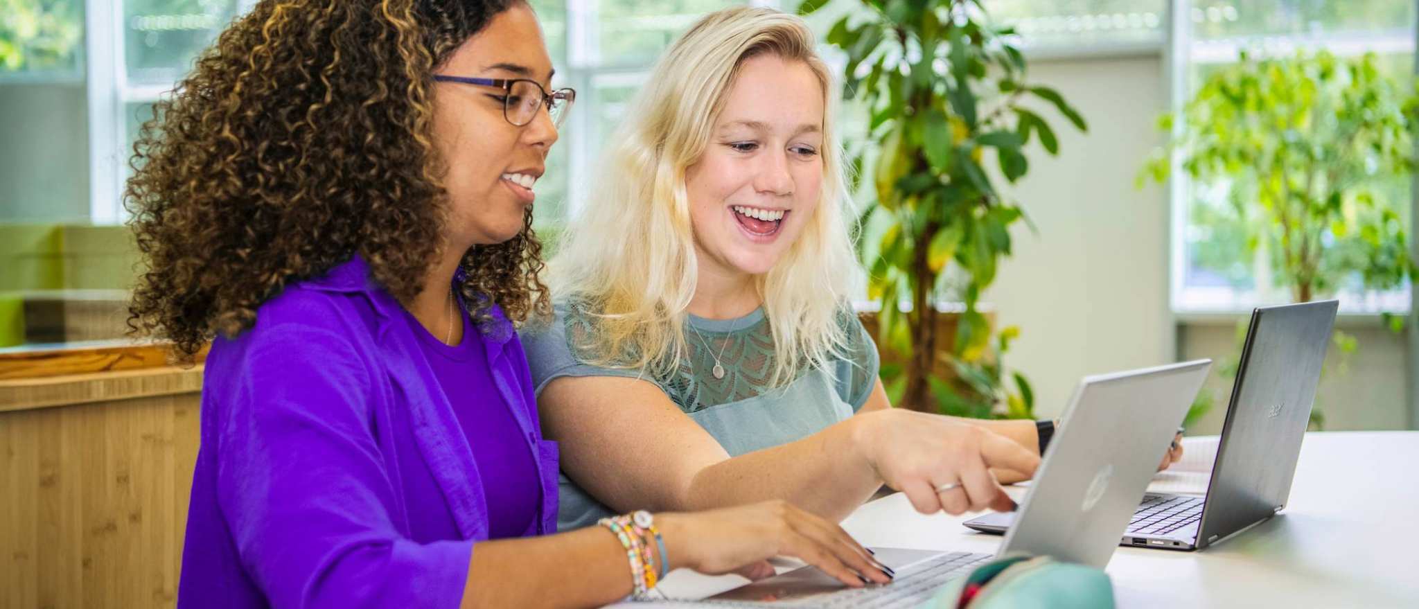 Twee studenten Leraar Wiskunde zitten naast elkaar met een laptop. De ene wijst lachend op het scherm iets aan en de ander kijkt lachend mee.