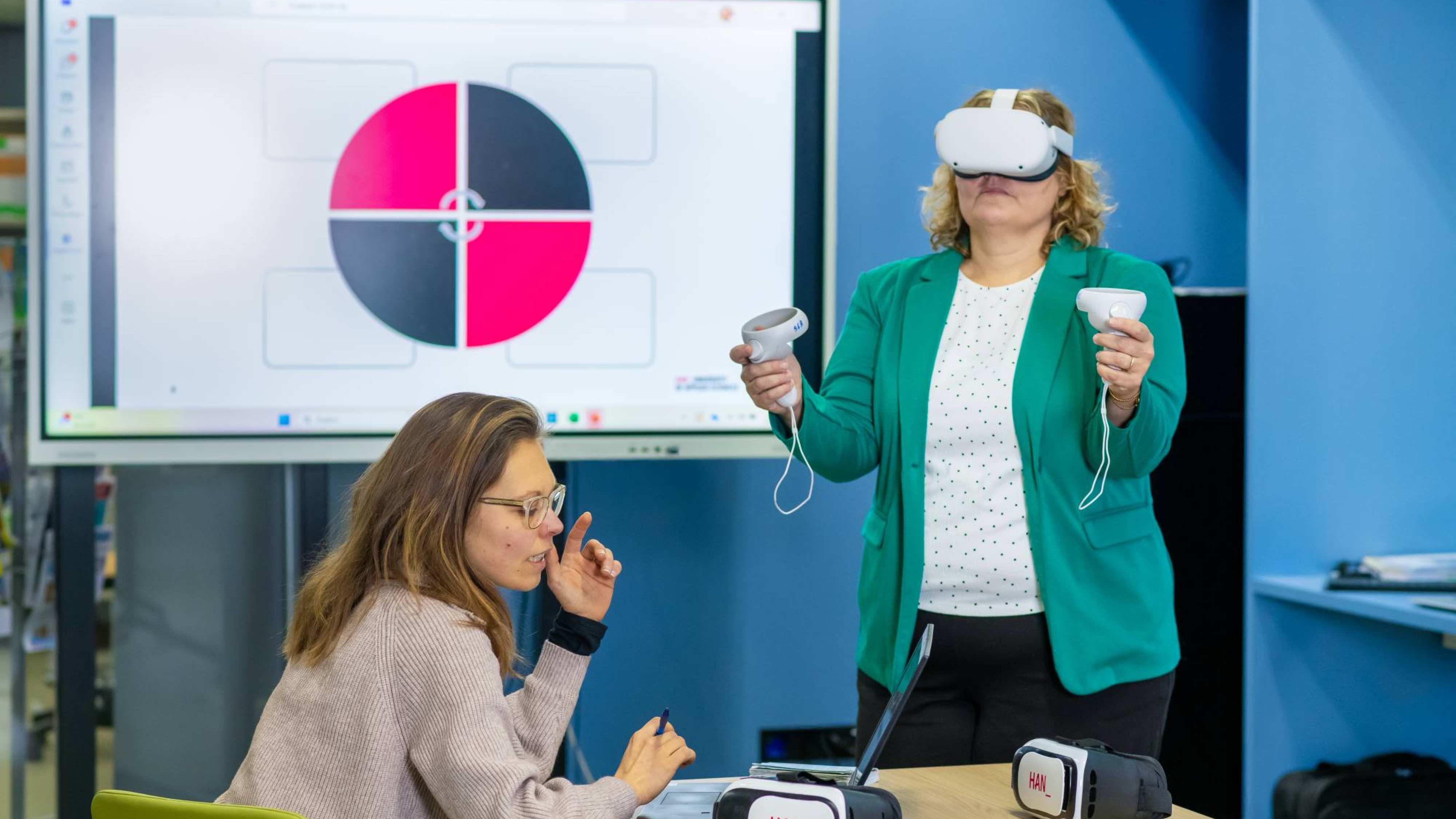 Iemand staat met een VR bril op en er zit iemand aan tafel, met op de achtergrond een scherm.