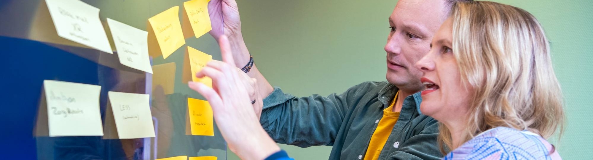 blonde vrouw met blauw en roze vest en man kort haar en groene blouse met geel tshirt, hangen gele post-its op een glazen wand