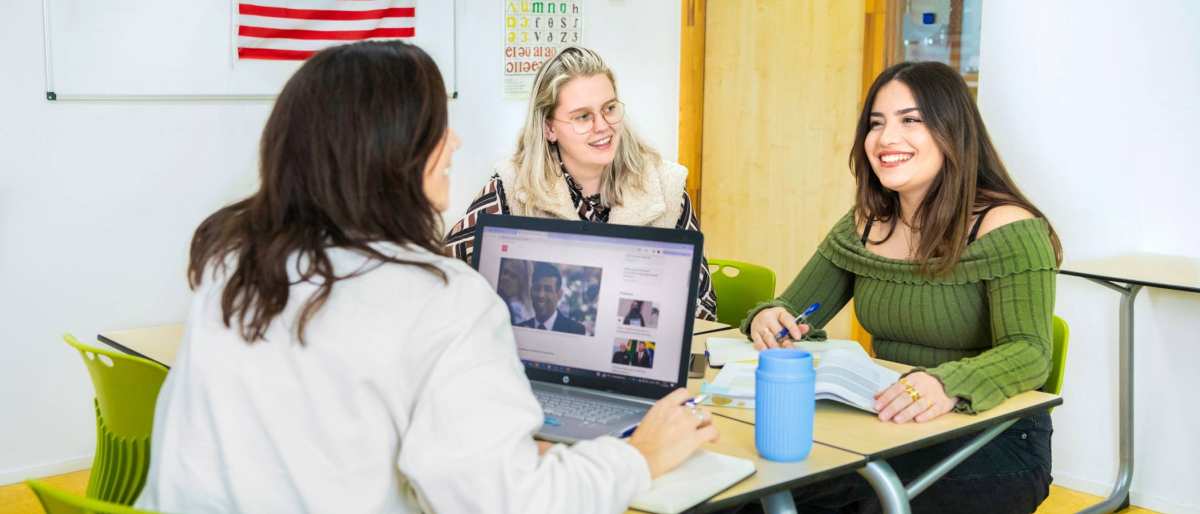 Drie studenten zitten in een Engels lokaal waar de Amerikaanse vlag op de achtergrond hangt