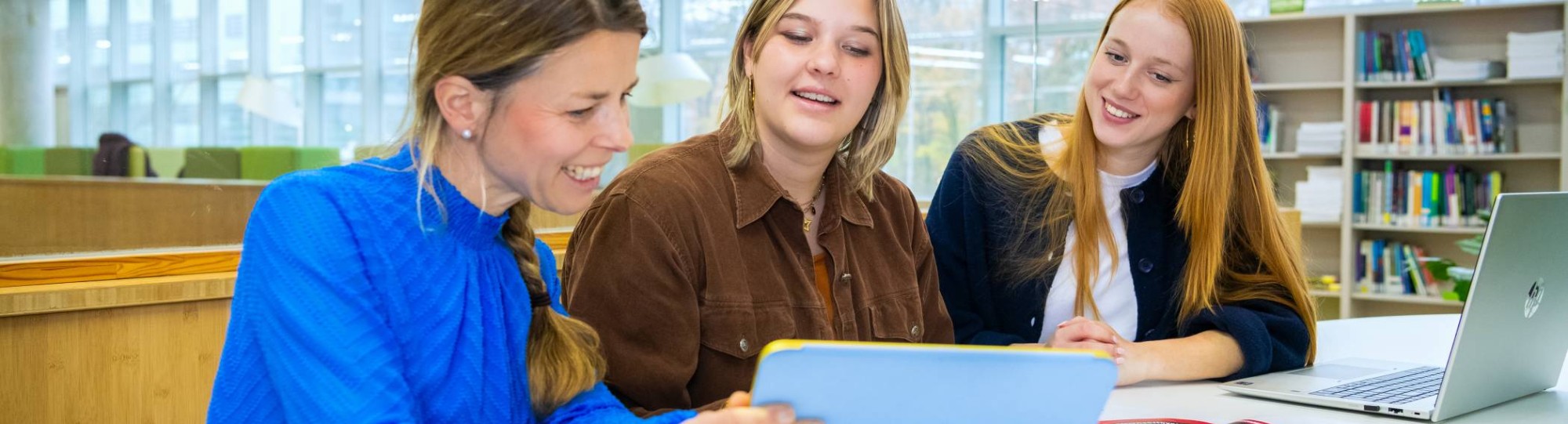 Drie mensen zitten aan een tafel met een tablet en een laptop en lachen.