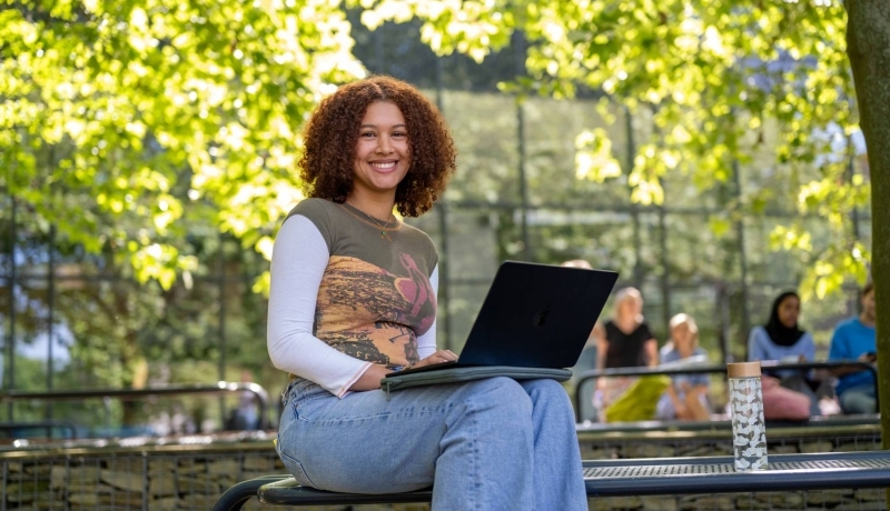 Studente kijkt in de camera, met laptop op schoot, zittend op de tafel, buiten bij K33. 