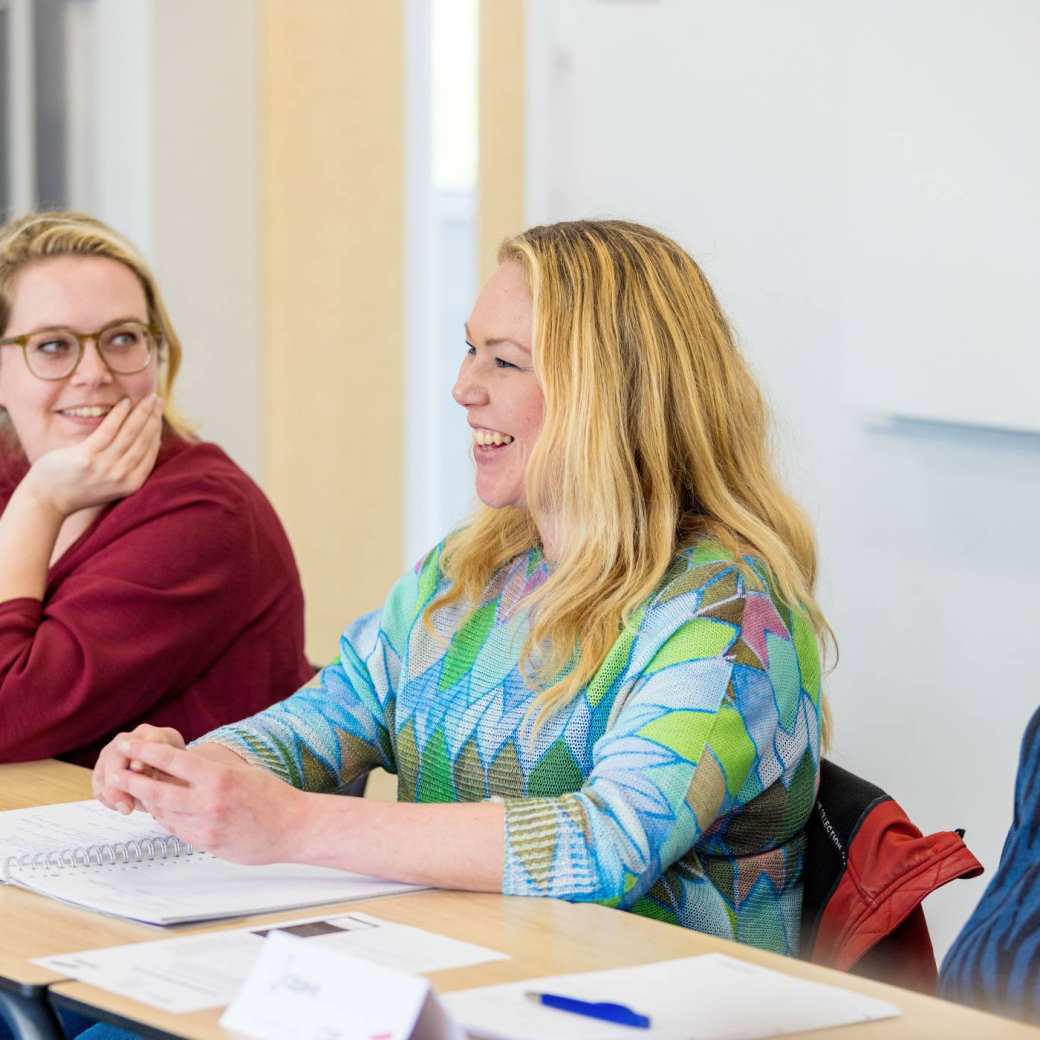 Vrouw is in gesprek en aan het lachen tijdens de cursus acute pijn service. Andere vrouw kijkt haar aan op de achtergrond.