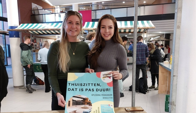 Hanne Wanink heeft blond haar en een groene top en draagt een gouden ketting. Romy Burghouts heeft een grijze jurk aan en donkere haren. Ze houden een poster in hun hand van het project met daarop Thuiszitten, dat is pas duur.