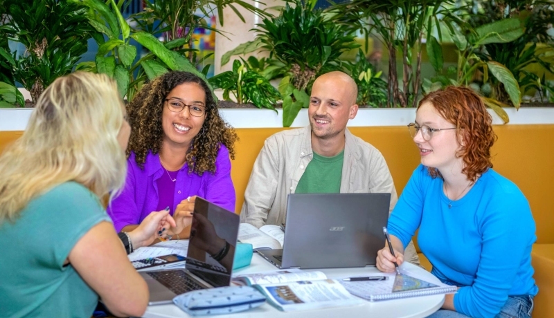 Vier studenten Leraar Wiskunde zitten aan een ronde tafel te werken. Er liggen laptops, etuis en schriften op de tafel.