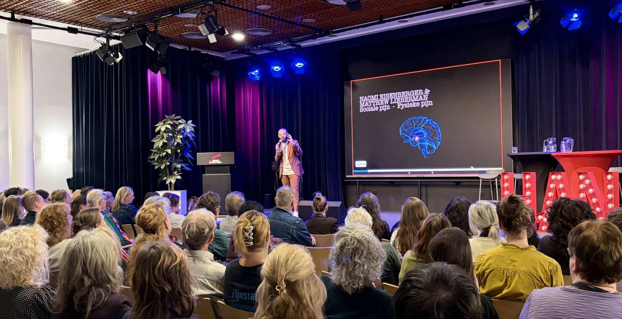 Volle zaal in Nijmegen met mensen in stoelen met op het podium schrijver/programmamaker Karim Amghar