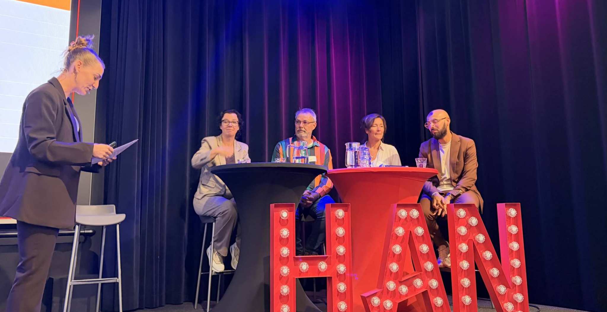 Op het podium zitten mensen achter een statafel. Er staan grote lichtgevende letters op het podium die HAN spellen