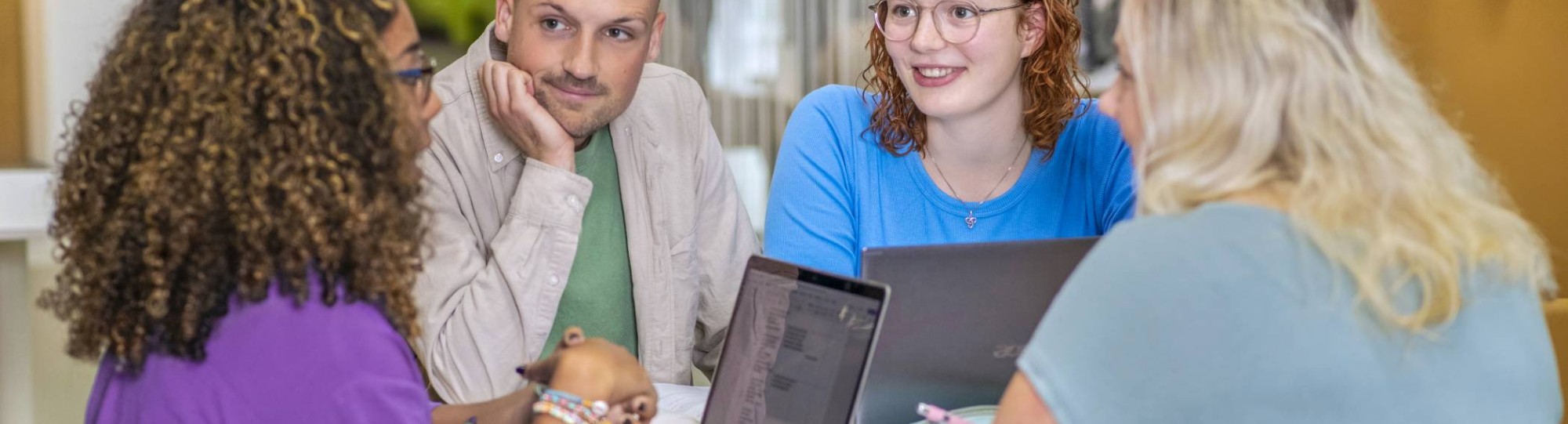 Vier studenten Leraar Wiskunde overleggen aan een ronde tafel en luisteren aandachtig naar elkaar. Op de tafel liggen etuis, schriften en laptops.