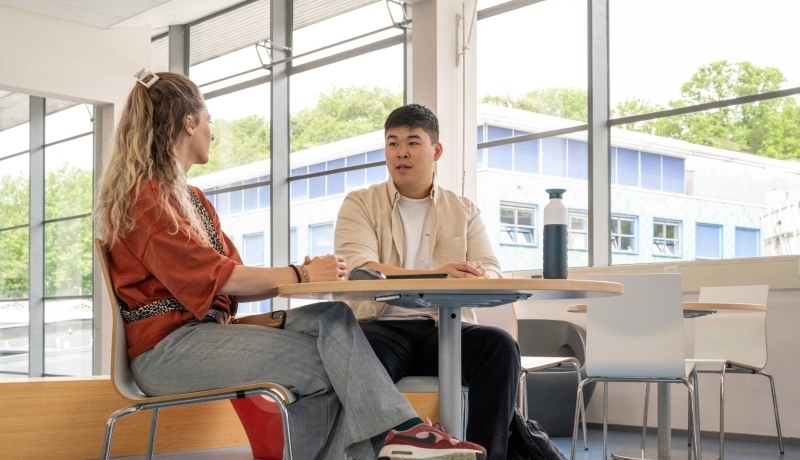 Student in gesprek met docent aan een tafel op podium in hal van gebouw R26, studentbegeleiding. 