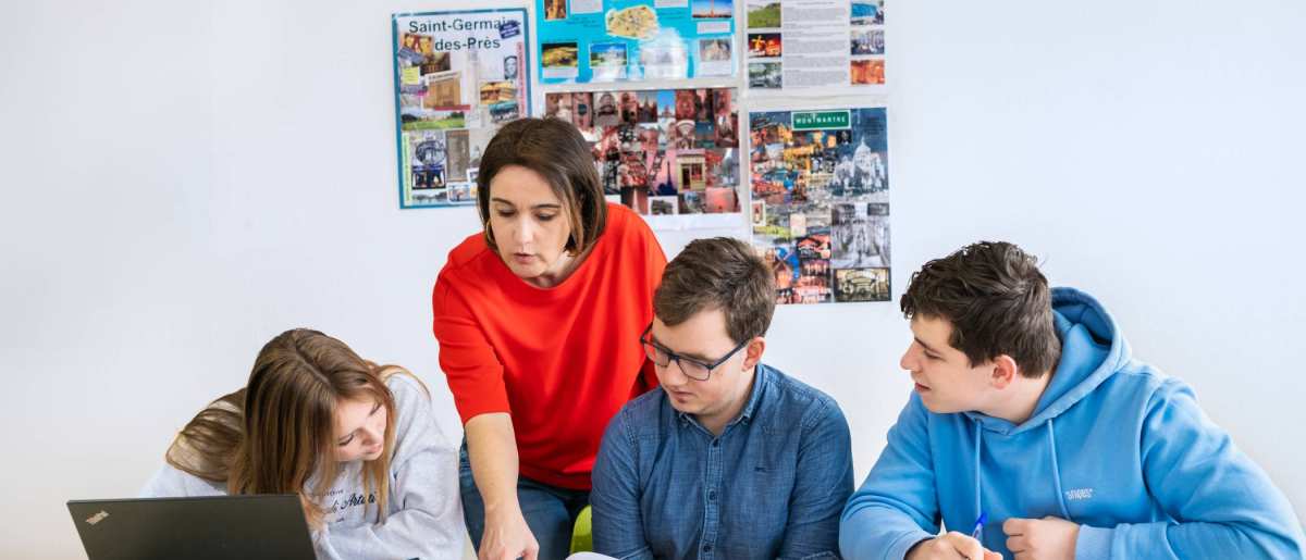 Drie studenten zitten op een rij en de docent staat achter hen. Ze wijst in het boek en de studenten kijken mee.