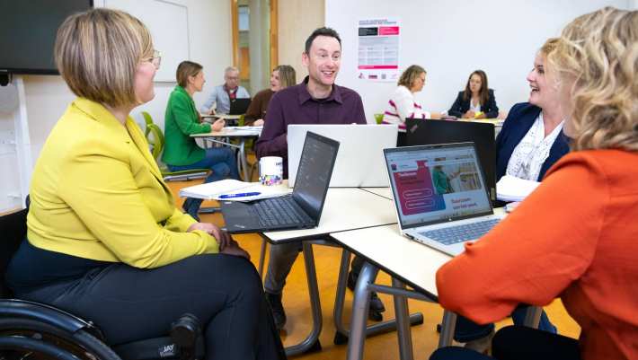 Een groepje van vier mensen in gesprek met laptops op tafel in een lokaal met anderen