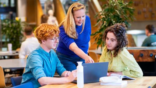 De lerares wijst naar de laptop waar twee studenten bij zitten.