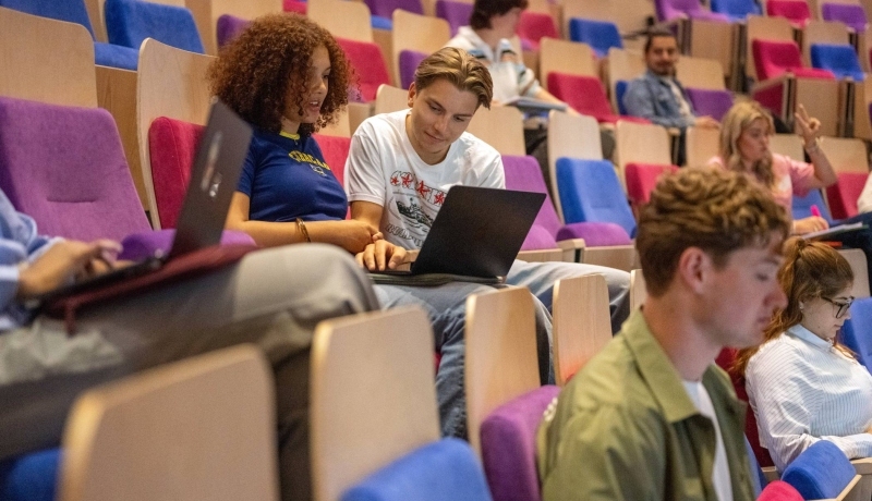 Twee studenten kijken samen op de laptop en overleggen in de collegezaal.