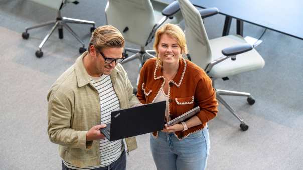 Masterstudent in bruin jasje op de HAN kijkt in camera, een andere masterstudent op zijn laptop.