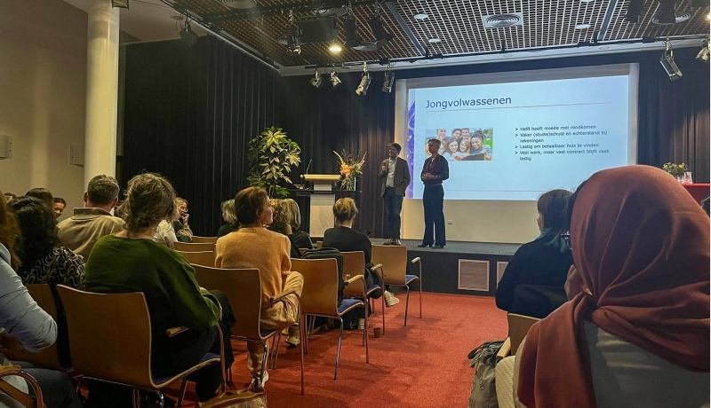 sfeerbeeld van de opening van de week tegen armoede. Mensen zitten op een stoel in een zaal met rode vloerbedekking. In het midden op de foto een podium met daarop Arjan Vliegenthart en Carinda Jansen