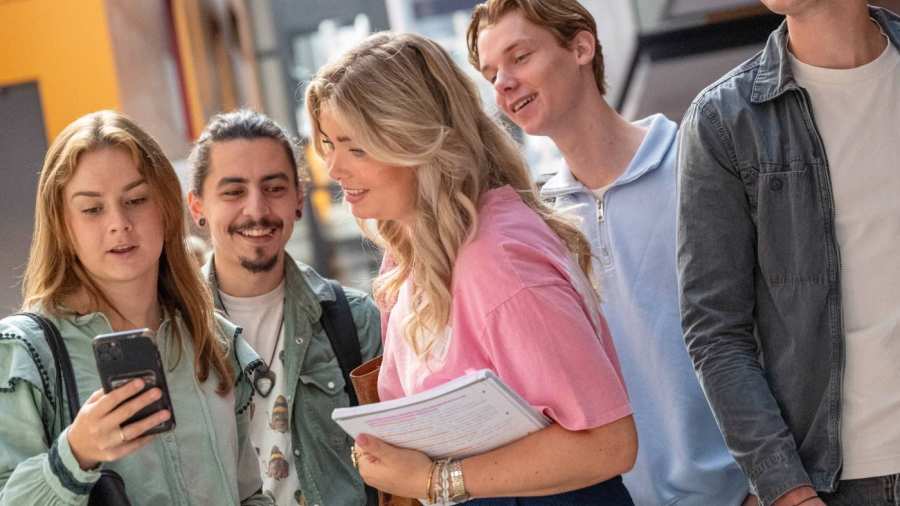 Groepje studenten staan bij elkaar en hebben plezier. Studenten kijkt op haar telefoon, ze staan in de gang.