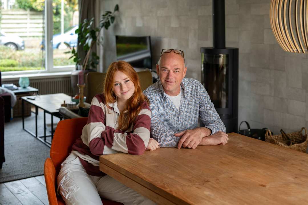 Vader en dochter kijken lachend in de camera, zitten aan tafel. 