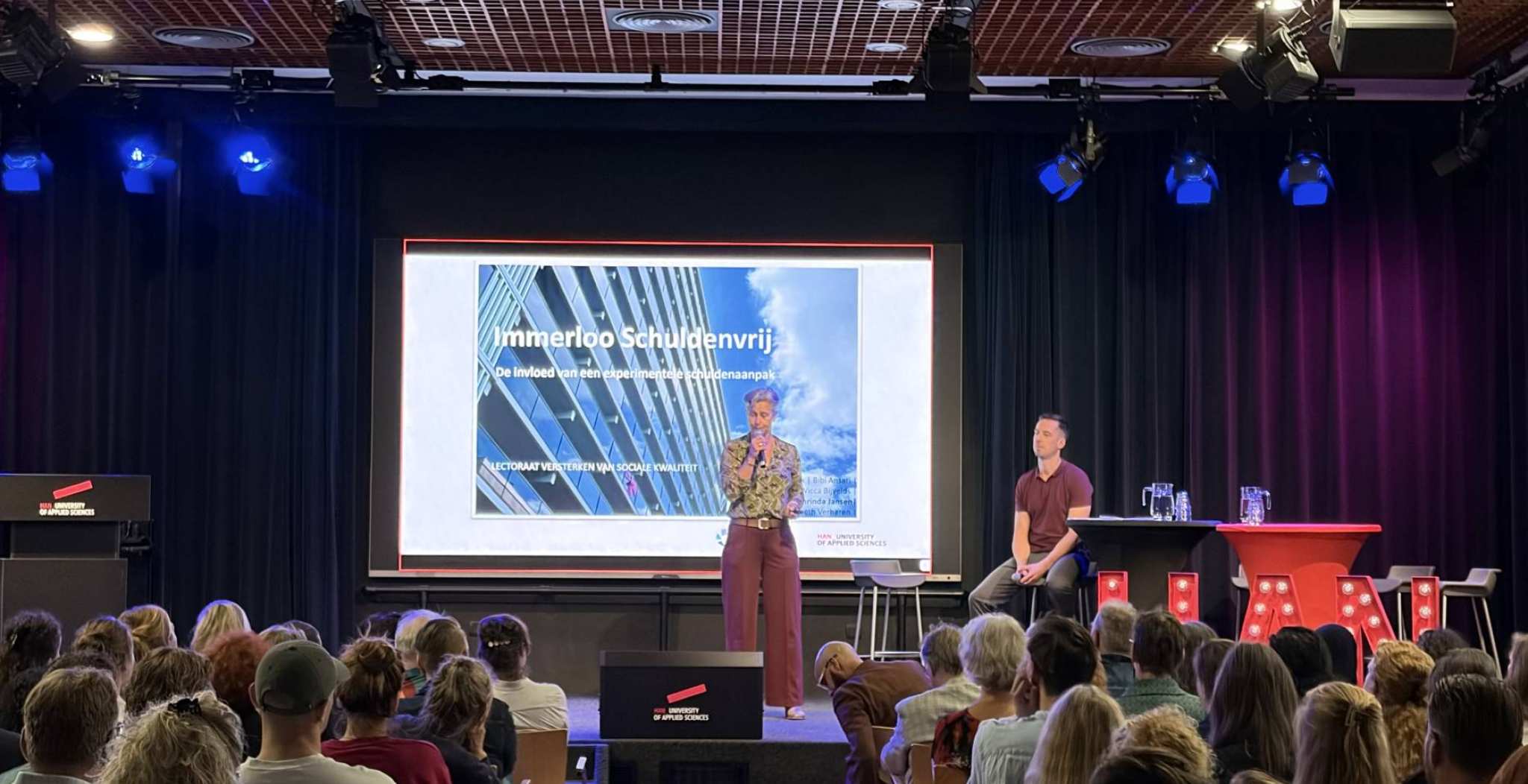 Op het podium staat een vrouw met een kort blonde kapsel te praten voor een scherm met daarop Immerloo Schuldenvrij