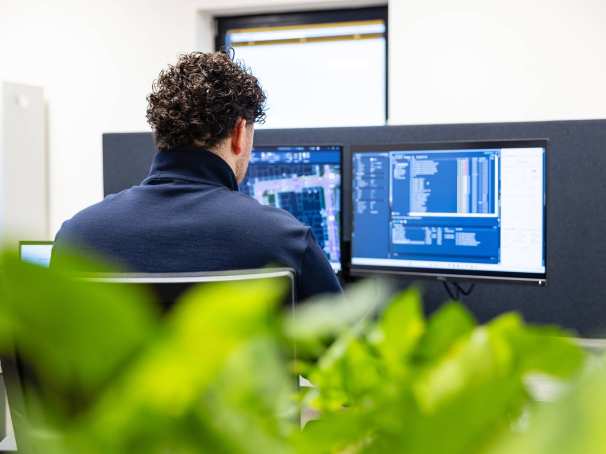 Man werkt op twee beeldschermen aan een bouwontwerp. Op de voorgrond staat een plant