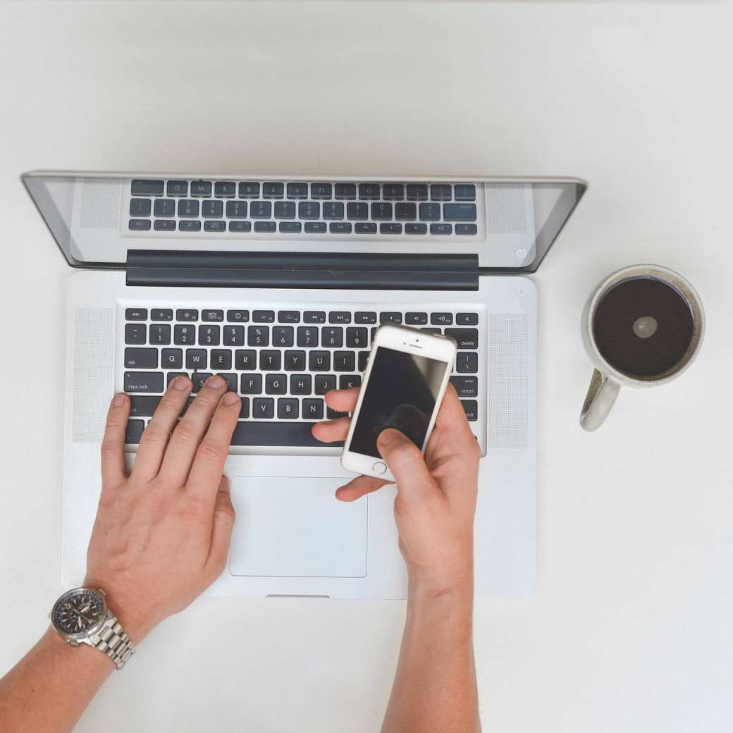 handen aan laptop met smartphone en kop koffie binnen handbereik. Macbook. iPhone