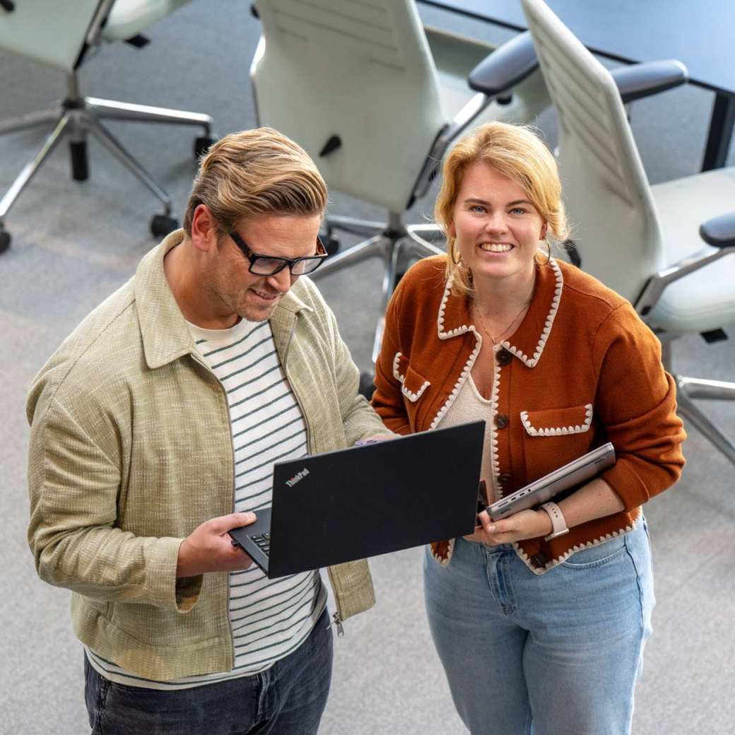 Man en vrouw staan met laptop in de hand en kijken lachend naar camera