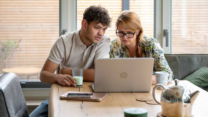 Moeder en zoon serieus bezig op de laptop, zittend aan tafel. 