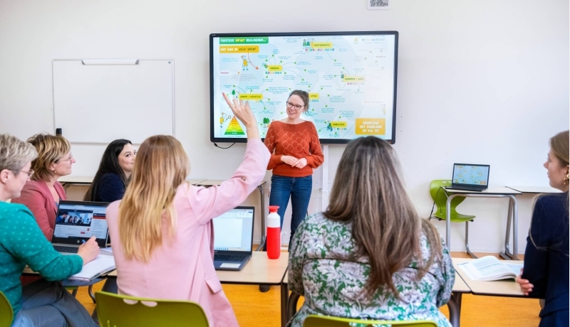 Docent staat voor een volle klas en kijkt lachend naar de student die de vinger opsteekt.