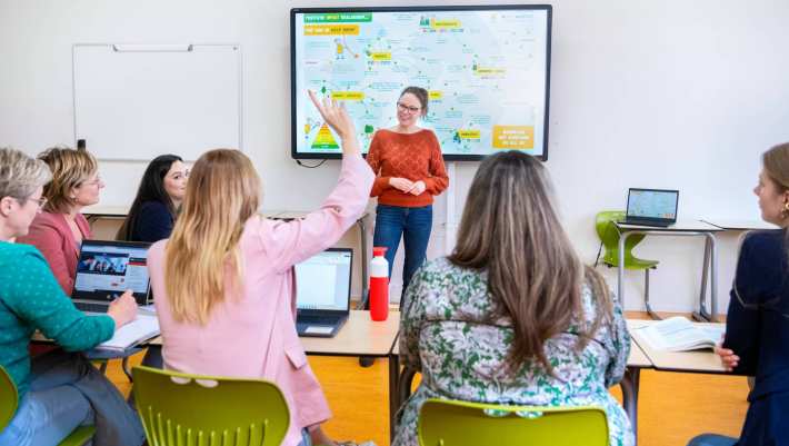 Docent staat voor een volle klas en kijkt lachend naar de student die de vinger opsteekt.
