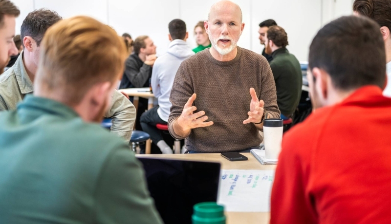 Docent zit bij studenten aan tafel en is aan het woord.
