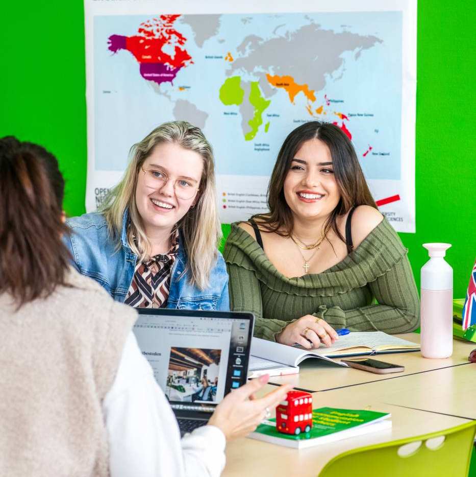 Twee studenten zitten naast elkaar en lachen naar de student die tegenover hun zit. Achter de twee studenten hangt een kaart met 'English Speaking Countries'