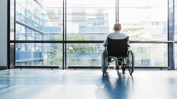 oudere man in rolstoel kijkt uit raam in een het ziekenhuisgang
