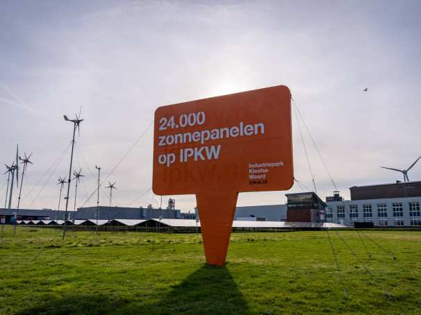 Foto van het Industriepark Kleefse Waard (IPKW) bij de ingang van het park met een bord over zonnepanelen capaciteit van het IPKW en met windmolens op de achtergrond. Academie Engineering en Automotive.