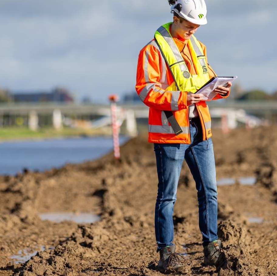 Man met veiligheidshelm en oranje werkjas checkt papieren op nog kale bouwgrond naast een kanaal.