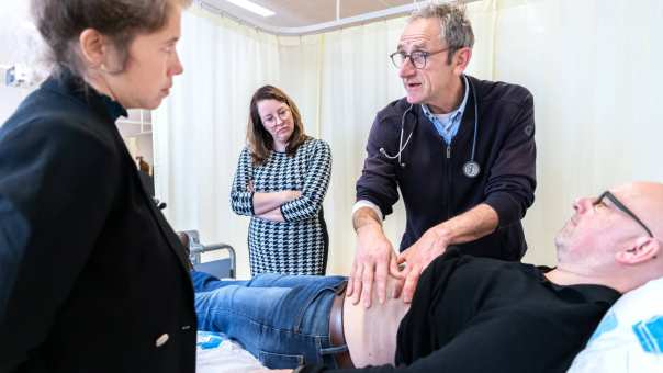 Docent van de HAN Master Physician Assistant geeft uitleg aan 2 studenten over palpatie bij een patiënt op de onderzoeksbank