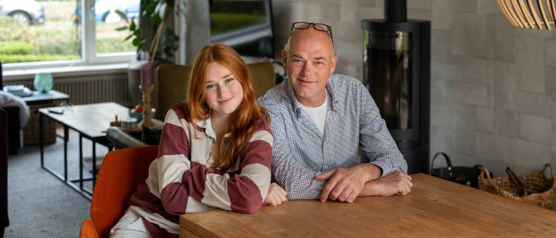 Vader en dochter kijken lachend in de camera, zitten aan tafel.