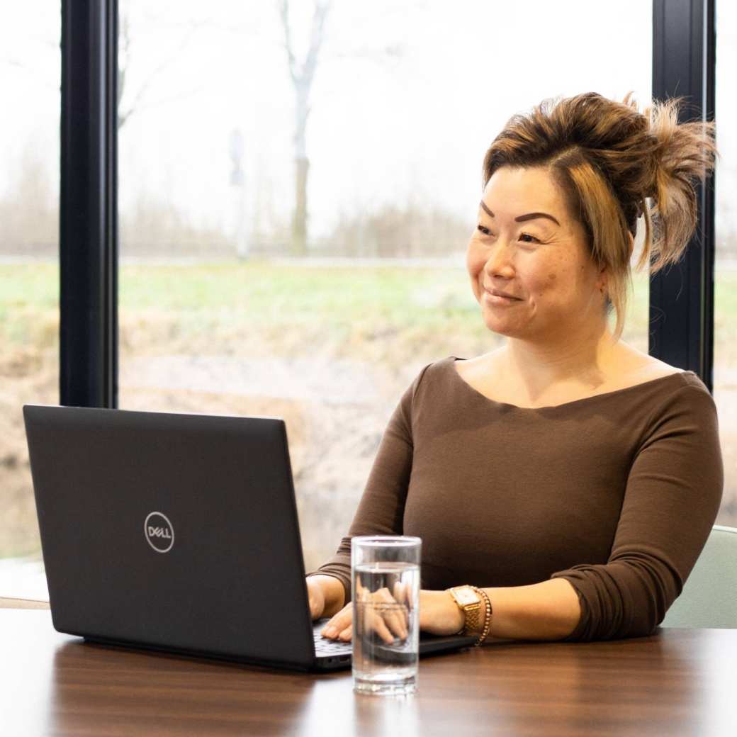 Dame poseert met een glimlach, achter een laptop. Een glas water staat naast haar op het bureau.