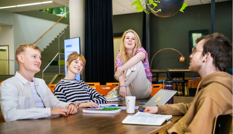 Drie studenten zitten om de tafel en een student zit op de tafel. Ze hebben schriften voor zich en praten met elkaar.