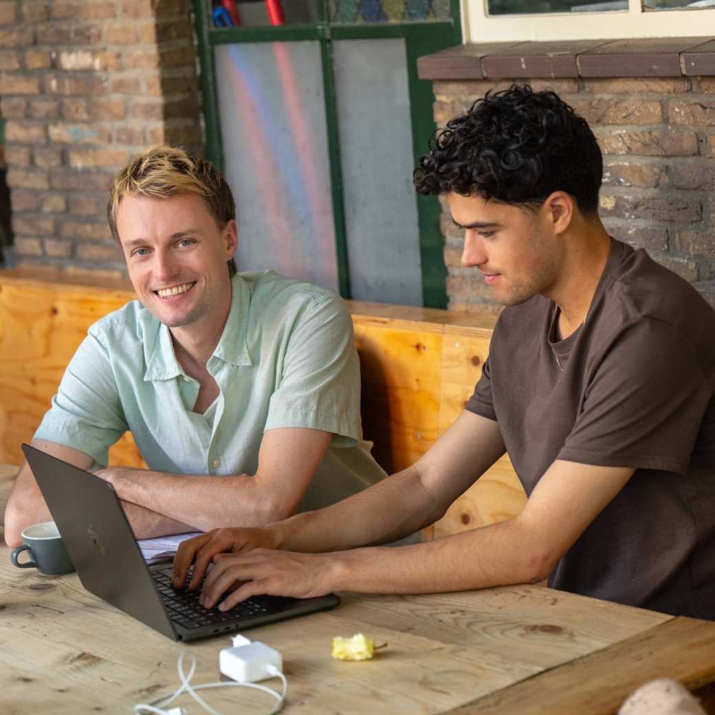 Twee studenten zitten aan tafel te studeren achter de laptop en lacht naar camera