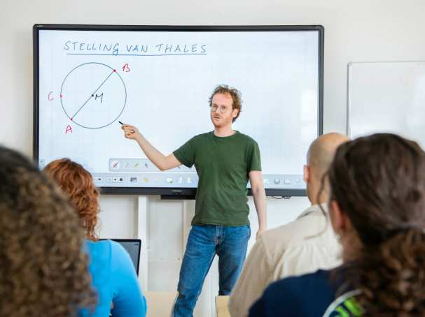Leraar Wiskunde kijkt naar zijn klas, terwijl zijn hand wijst naar de cirkel op het bord waar 'stelling van Thales' boven staat