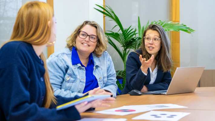 Drie mensen aan tafel in gesprek met elkaar, met een hoop papieren op tafel en een laptop